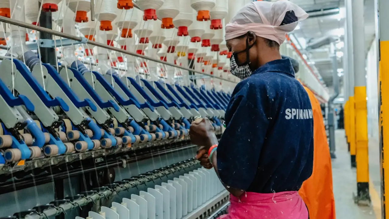 A textile worker in a headscarf and face mask operates a spinning frame, with rows of yarn bobbins overhead, at a factory.