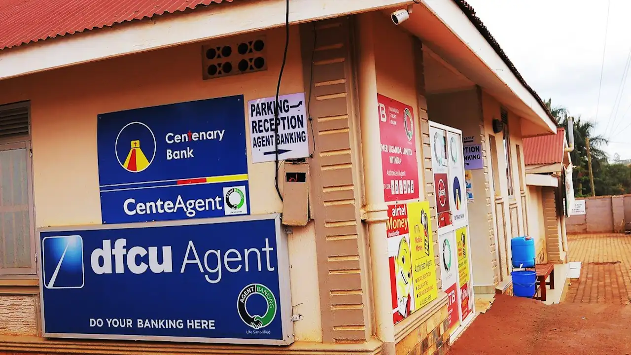 Single-storey building with a red roof and signs for Centenary Bank's CenteAgent and dfcu Agent, plus Airtel Money and other agent notices by the door.