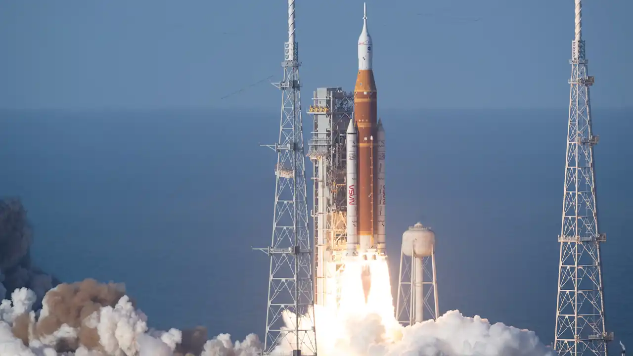 Artemis II launch | Uganda Business News A wide shot of the Artemis SLS rocket launching from Kennedy Space Center. The orange core stage and twin white solid rocket boosters emit a bright white glare and thick clouds of exhaust. Three tall metal lightning towers frame the launch pad against a clear, pale blue horizon.