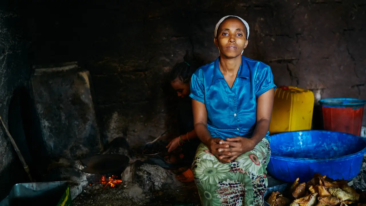 A woman sits in a kitchen while someone else tends to a wood-fired, open, three-stone stove behind her.