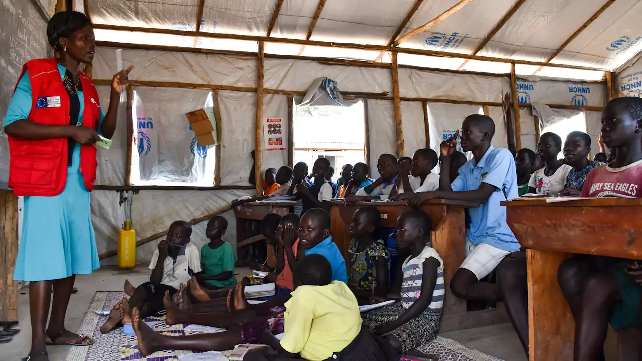 Children attending school in a refugee settlement in northwestern Uganda in 2019. Some children are sitting on a mat at the front of the class, while others are seated at wooden desks. A female teacher is leading the crowded class.