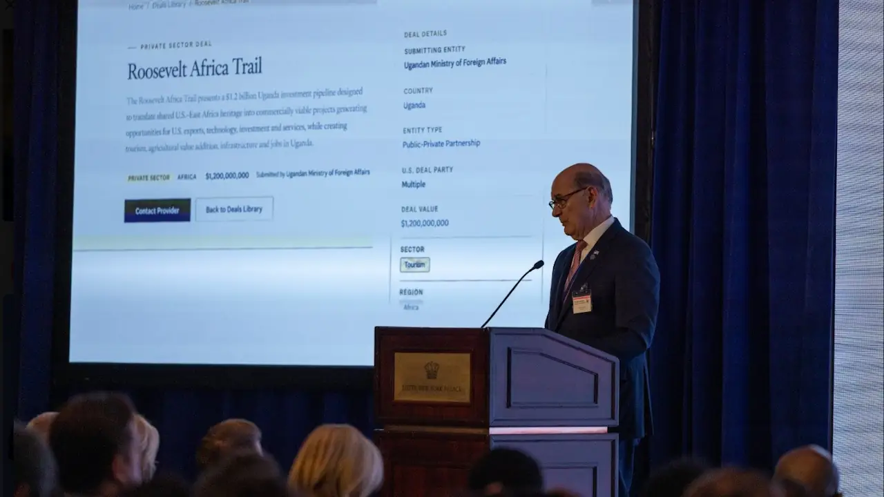 Ambassador Dan Negrea speaks at a lectern in front of a large screen showing the Roosevelt Africa Trail listing on the US State Department's Trade Over Aid Deals Library website, with a deal value of $1,200,000,000 for Uganda