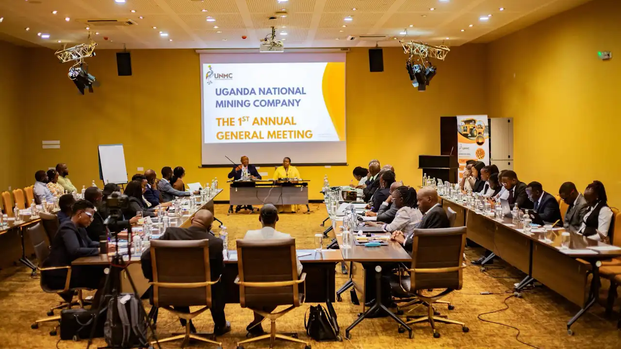 Delegates seated at long tables arranged in a U-shape in a yellow-walled conference room, facing a head table where Ruth Nankabirwa, in yellow, sits beside a man in a dark suit beneath a screen reading "Uganda National Mining Company: The 1st Annual General Meeting"