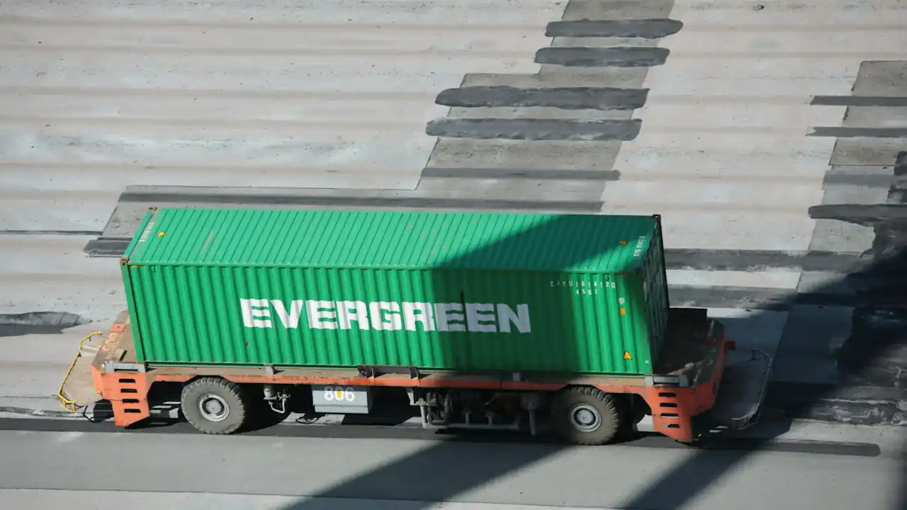 An aerial view of a bright green Evergreen shipping container being transported on a red automated guided vehicle across a paved port terminal.