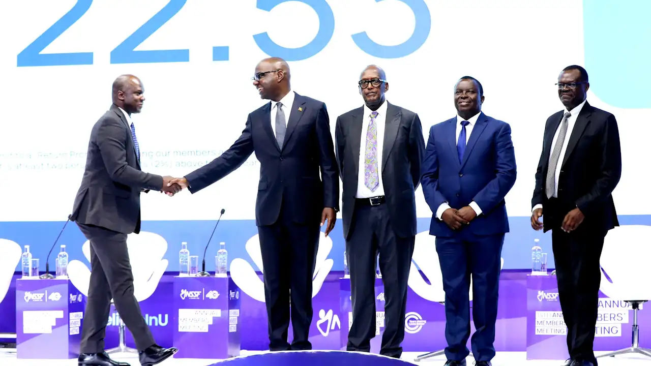 Five men in dark suits stand on a stage at NSSF's annual members' meeting. Finance minister Henry Musasizi, second from left, shakes hands with a man on the far left, beside gender minister Henry Tumukunde, centre, and NSSF managing director Patrick Ayota, far right. The figure 22.53 is displayed on a large screen behind them.