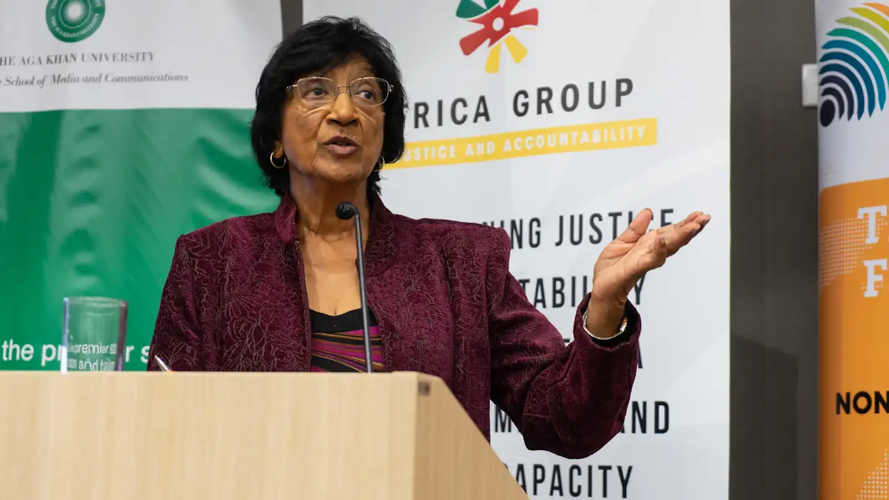 Navi Pillay, in a burgundy jacket and glasses, speaks at a lectern and gestures with one hand, in front of banners for the Africa Group for Justice and Accountability.