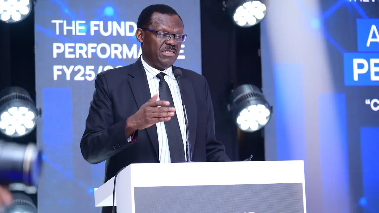 Patrick Ayota, NSSF managing director, gesturing while speaking at a lectern, with a screen behind him reading "The Fund Performance FY25/26"