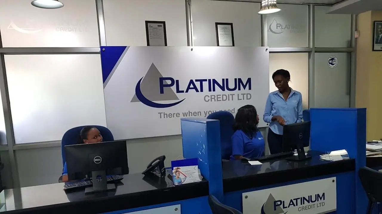 Reception area of a Platinum Credit Ltd branch, with two staff members seated at computers behind blue counters and a third in a blue shirt standing between them, below a wall sign bearing the company's logo and tagline