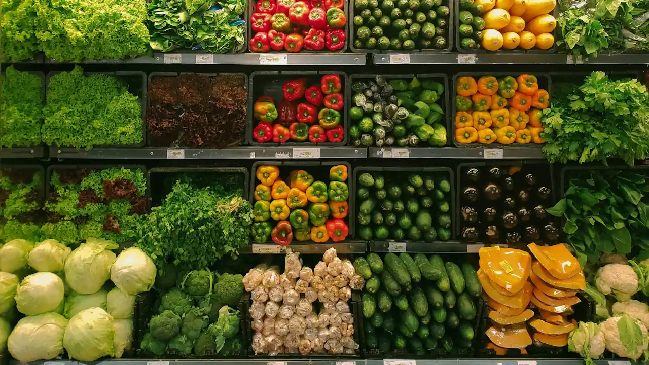 Organised supermarket shelves displaying a variety of fresh vegetables including green peppers, cabbages, pumpkins, greens, and brassicas.