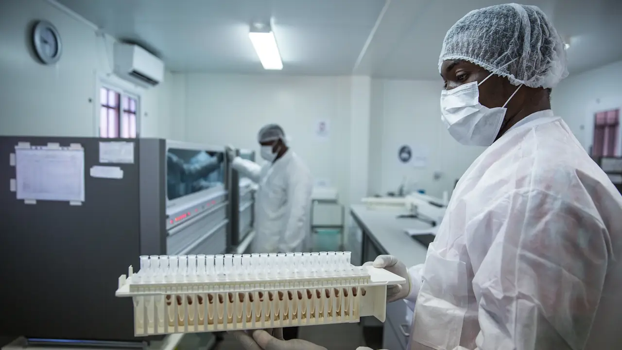 Viral load testing in Mozambique | Uganda Business News A laboratory technician in white protective gear and a face mask holds a rack of test tubes in a bright, modern molecular biology lab in Mozambique. In the background, another technician works at a large diagnostic machine.